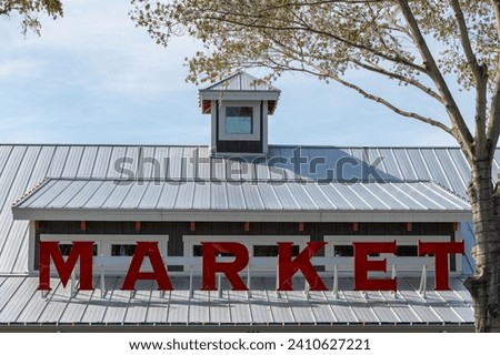 A vibrant red illuminated market sign on the roof of a rustic building. The storefront has a peaked roof, large window and wooden exterior wall. The background is blue sky with clouds and green trees.
