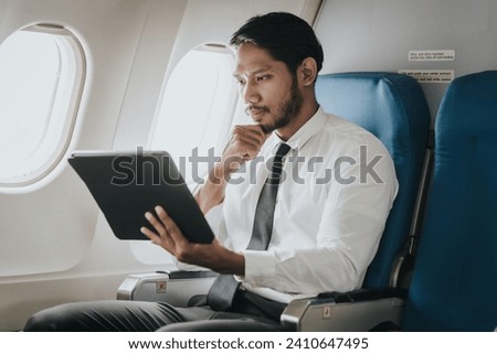 Asian businessman, possibly a stock market trader or investor, focused on his tablet while flying, indicative of a professional managing work on the go.