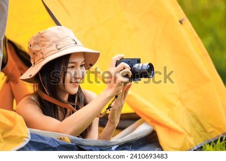 Young asian female tourist in safari dress and camera in her hands while lying inside the tent in the midst of nature in the morning,
