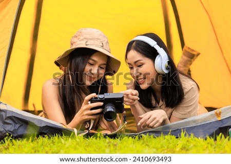 two young asian female tourist in safari dress and camera in her hands with headphone music while lying inside the tent in the midst of nature in the morning,
