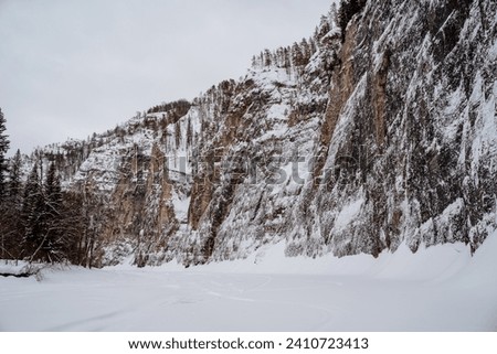 A beautiful mighty rock on the bank of a frozen river in winter, a beautiful winter landscape, cloudy weather in the mountains, a taiga forest. High quality photo