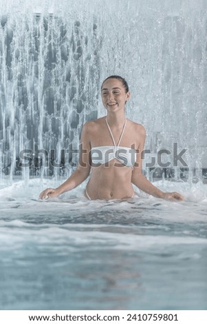 Optimistic young female in white bikini smiling and looking away while standing near wall of falling water during hydrotherapy session on weekend day on spa resort Royalty-Free Stock Photo #2410759801