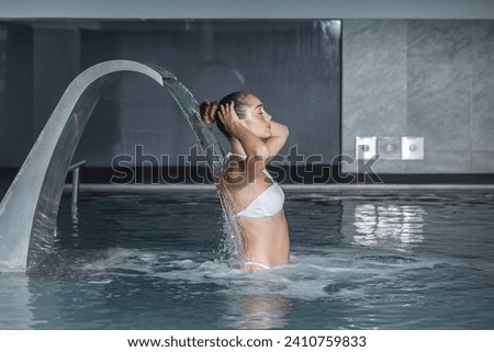 Side view of young female in bikini closing eyes and touching hair while standing in clean pool under fountain during spa session on resort Royalty-Free Stock Photo #2410759833