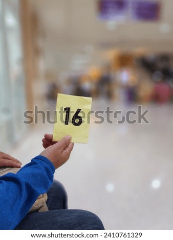 A yellow queue card printed with black letters indicates the number 16. A right hand holds a large paper queue card with the number 16 while waiting at hopital for service on a blurred background.