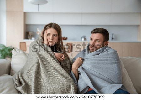 Unhealthy woman with headache sit on couch near unpleased husband wrapped in blanket from insufficient heating, feeling unwell. Sick couple at home suffering from cold. Freezing during heating season Royalty-Free Stock Photo #2410770859
