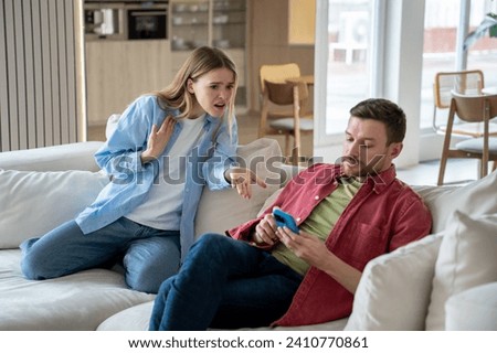 Indifferent ignoring man sits on sofa with mobile phone, playing games, scrolling social networks, reading news, keeping silence while offended irritated woman raises voice and shouts reproachfully Royalty-Free Stock Photo #2410770861