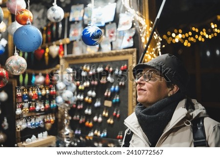 Old woman shopping at the Christmas street market, choosing gifts. Winter holidays, travel, shopping conception. Evening portrait in the open air.