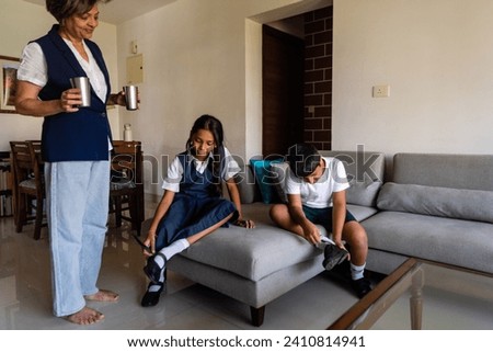 A tired young boy and his pre-teen sister sit on the couch and take off their shoes after coming home from school as their grandmother hands them some water to drink in steel glasses in Mumbai, India.