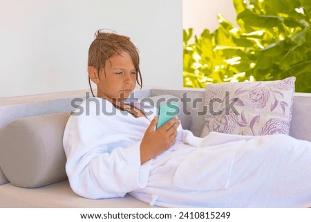 cute teenage boy in a white bathrobe lies on a sunbed on the ocean in the Maldives and surfs on her phone