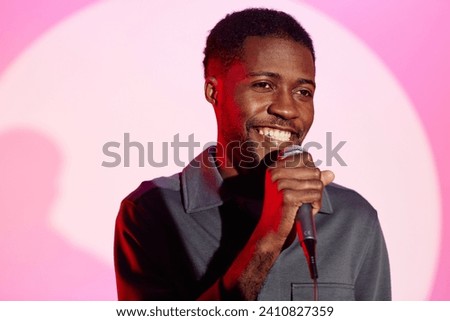 Medium shot of cheerful African American young man at light pink background speaking into microphone at party while looking away