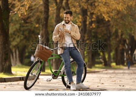 Young man with bicycle using smartphone in autumn park, space for text