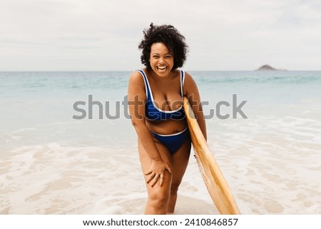 Young female surfer having fun on a beach adventure, holding her surfboard and wearing a vibrant bikini. Curvy woman smiling at camera, enjoying an active lifestyle and solo summer fun. Royalty-Free Stock Photo #2410846857