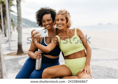 Young women enjoy a beach workout, taking selfies and capturing a happy, healthy lifestyle on camera. Two fitness friends enjoying a wellness day with ocean views and beach vibes.