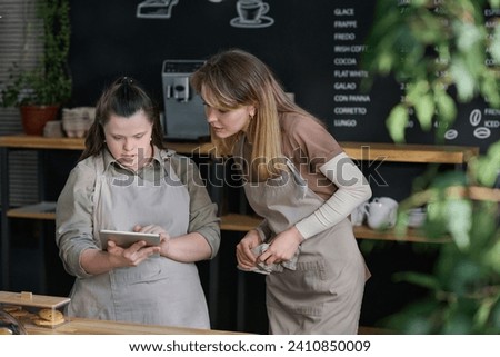 Young waitress with Down syndrome showing something in tablet to colleague during discussion of working points by counter Royalty-Free Stock Photo #2410850009