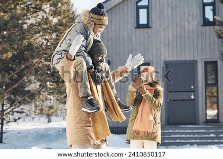 Happy young man in winterwear having fun with cheerful cute boy sitting on his neck while woman with smartphone taking picture of them