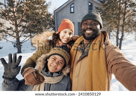 Happy young family of three taking selfie and looking at camera while standing against their country house or cottage on winter day
