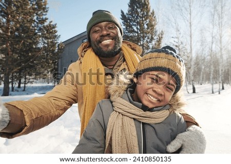 Cheerful African American father and son in winterwear looking at camera in rural environment on frosty winter day while taking selfie