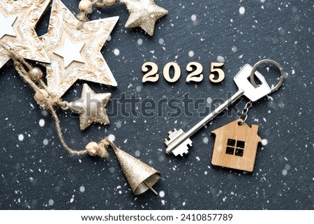 House key with keychain cottage on festive black background with stars, lights of garlands. New Year 2025 wooden letters, greeting card. Purchase, construction, relocation, mortgage, insurance