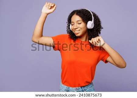 Little kid teen girl of African American ethnicity wear orange t-shirt listen to music in headphones dance raise up hands isolated on plain pastel purple background studio. Childhood lifestyle concept