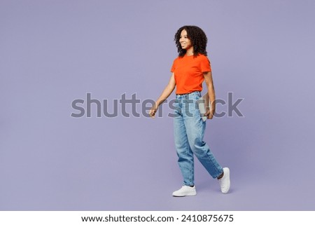 Full body side view little kid teen IT girl of African American ethnicity wear orange t-shirt hold closed laptop pc computer walk go isolated on plain purple background. Childhood lifestyle concept