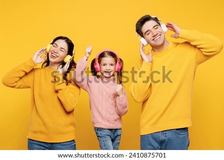 Young happy cool parents mom dad with child kid girl 7-8 years old wear pink sweater casual clothes listen to music in headphones dance have fun isolated on plain yellow background. Family day concept Royalty-Free Stock Photo #2410875701