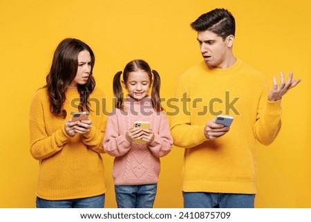 Young sad indignant parents mom dad with child kid girl 7-8 years old wearing pink sweater casual clothes hold use looking at mobile cell phone isolated on plain yellow background. Family day concept Royalty-Free Stock Photo #2410875709