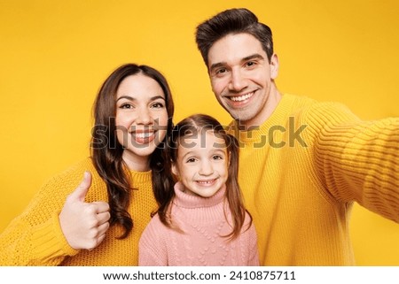 Close up young parents mom dad with child kid girl 7-8 years old wear pink sweater casual clothes do selfie shot mobile cell phone show thumb up isolated on plain yellow background. Family day concept