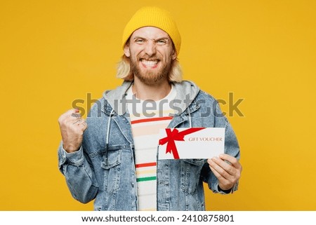 Young man wears denim shirt hoody beanie hat casual clothes hold gift certificate coupon voucher card for store do winner gesture isolated on plain yellow background studio portrait. Lifestyle concept