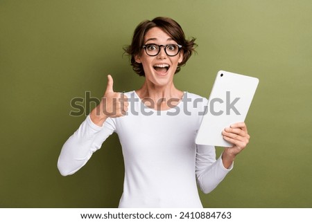 Photo portrait of attractive young woman hold tablet show excited thumb up dressed stylish white clothes isolated on khaki color background