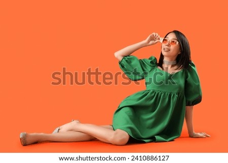Beautiful stylish Asian woman in sunglasses sitting on orange background