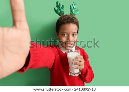 Little African-American boy in reindeer horns with glass of milk taking selfie on green background