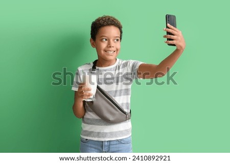 Little African-American boy with glass of milk taking selfie on green background