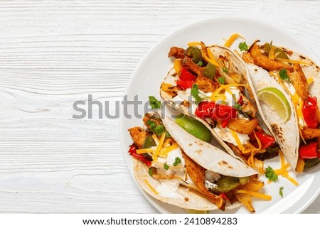 baked tex-mex chicken fajitas with mixed sweet pepper, onion, sour cream, cheddar cheese, lime, parsley and gluten free corn tortillas on plate on white wooden table, free space, flat lay, close-up