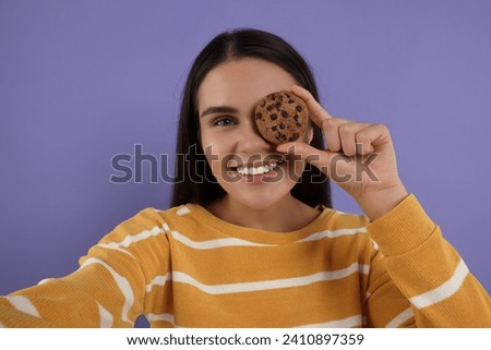 Young woman with chocolate chip cookie taking selfie on purple background