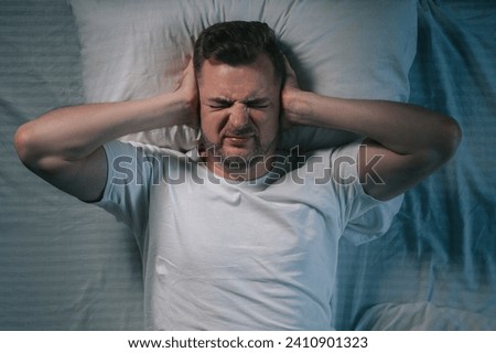 A young man covers his ears with his hands. The guy can't sleep in bed because of the loud noise. Top view. Royalty-Free Stock Photo #2410901323
