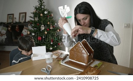 Mother creating gingerbread house with her son, applying royal ice. Family preparing for december holidays at home