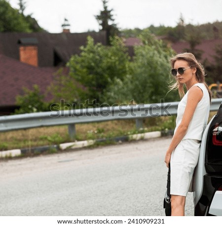 A fashionable beautiful slender blonde girl in sunglasses, light shorts and a vest, in a black leather handbag in her hands, stands on the track near a white car.