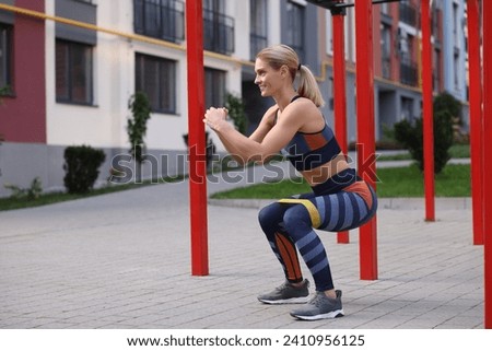 Athletic woman doing squats with fitness elastic band at outdoor gym. Space for text