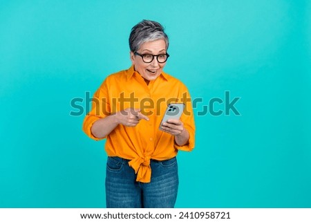 Photo of shocked mature aged retired woman point finger smartphone amazed low prices on amazon isolated over cyan color background