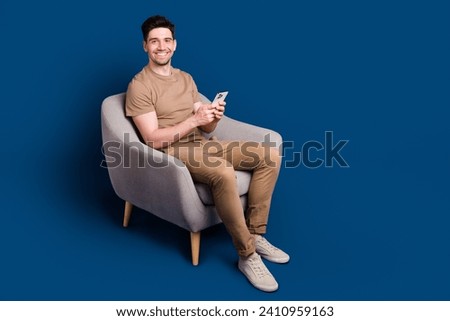 Full size photo of handsome man dressed beige t-shirt sit in armchair chatting on smartphone isolated on dark blue color background