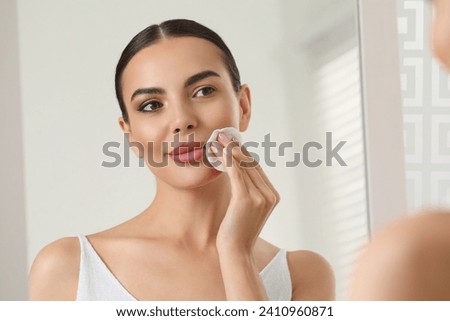 Beautiful woman removing makeup with cotton pad near mirror indoors Royalty-Free Stock Photo #2410960871