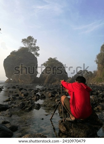 Unrecognizable photographer in red jacket takes a landscape picture of a beach with huge majestic rocks in the seashore. Beautiful view of a rocky beach in Pacitan, East Java, Indonesia