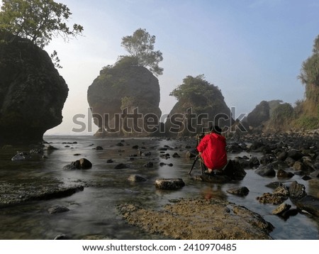 Unrecognizable photographer in red jacket takes a landscape picture of a beach with huge majestic rocks in the seashore. Beautiful view of a rocky beach in Pacitan, East Java, Indonesia