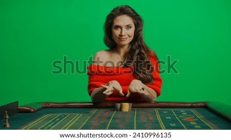Gorgeous female in studio on chroma key green screen. Appealing woman in red dress sitting at the roulette table smiling placing bet.