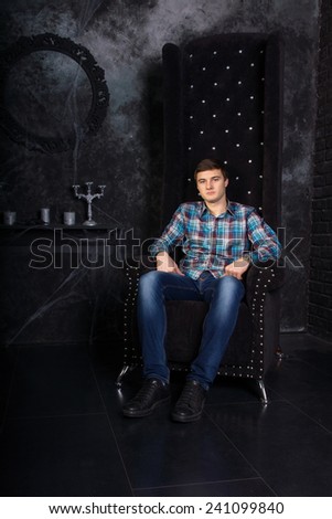 Man Wearing Casual Clothing Sitting in High Back Chair in Eerie Halloween Haunted House Setting with Candelabras and Cobwebs