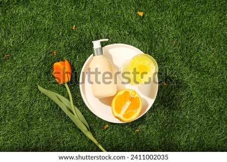Top view of a round white dish container a pump cosmetic bottle, half an orange and smoothie cup decorated with tulip flower on a green grass background. Top view, mockup bottle for design