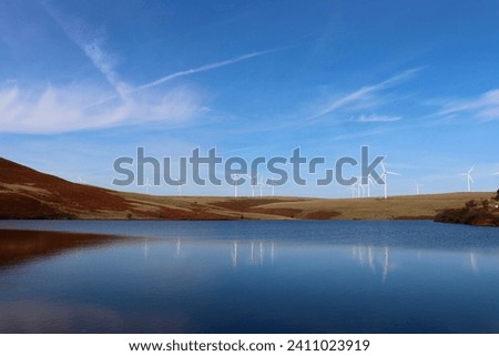 This image depicts a stunning outdoor landscape, with a body of water in the foreground and a group of wind turbines in the distance. The sky is filled with white clouds, creating a picturesque scene Royalty-Free Stock Photo #2411023919