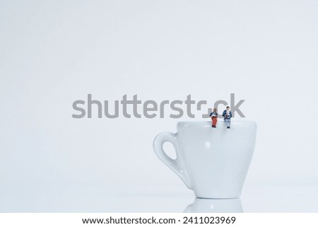 A couple sits reading on a white coffee cup, white background, miniature figure scene Royalty-Free Stock Photo #2411023969
