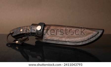 A hand-forged knife with a black horn handle in a leather sheath on black glass, which mirrors the knife