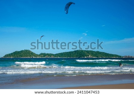 The picture shows two kite surfers in the ocean in front of a small, mountainous, forest-covered green island. Campeche Island, Ilha de Santa Catarina. 
